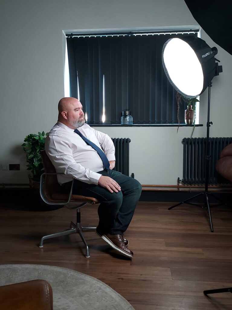 Image shows man in shirt and tie being filmed for a testimonial video. He's lit with professional video lights which reduce flickering on the footage