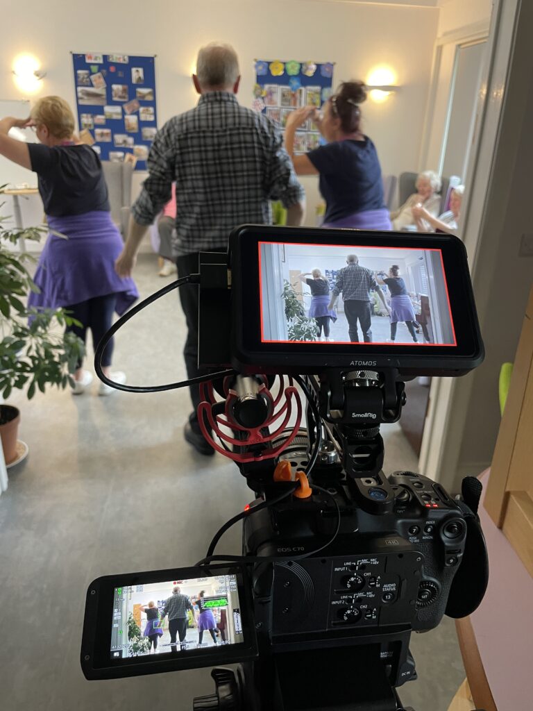 Photo shows video camera recording person living with dementia and two members of staff dancing together during activity session